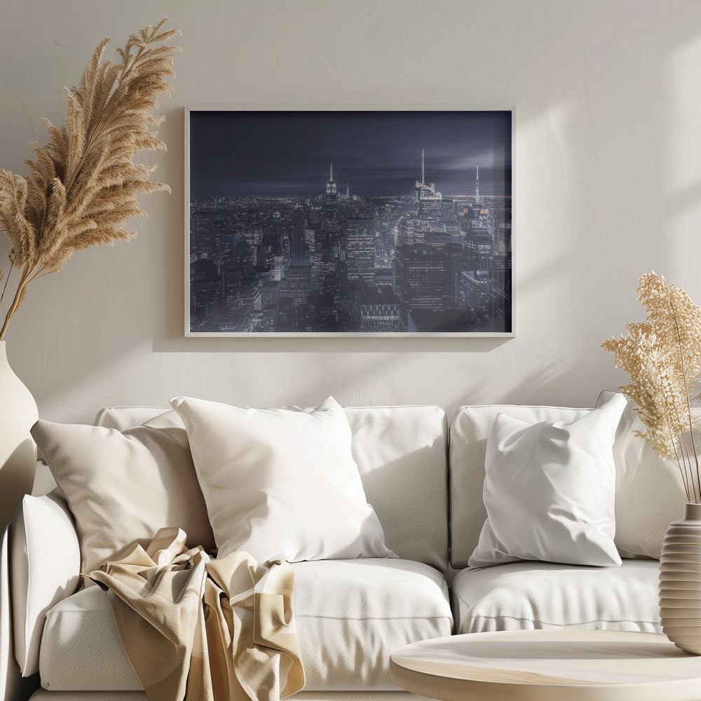 A dramatic nighttime photograph of the New York City skyline, viewed from a high vantage point. The cityscape is a sea of illuminated skyscrapers under a dark, moody sky, with the entire image cast in a cool blue and white monochrome tone and presented in a silver frame. Artwork