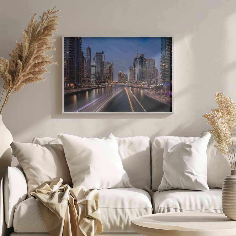A long-exposure photograph of the Dubai Marina at dusk, framed in silver. Illuminated skyscrapers line a busy waterway, with light trails from moving boats creating long, bright streaks on the water. The sky is a deep twilight blue, and the city's lights cast a warm glow. Decor