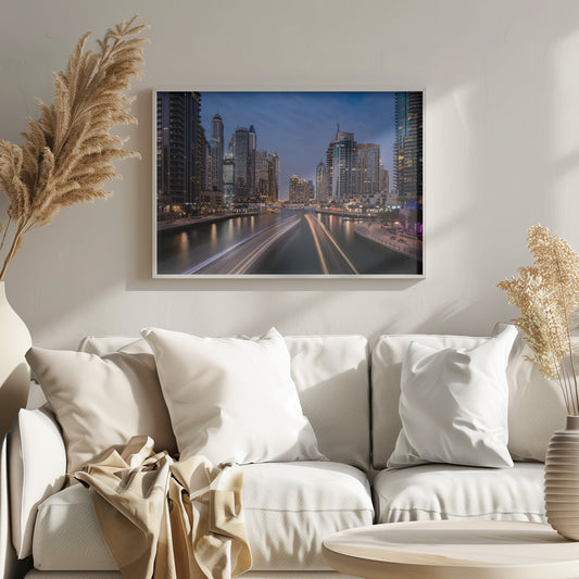 A long-exposure photograph of the Dubai Marina at dusk, framed in silver. Illuminated skyscrapers line a busy waterway, with light trails from moving boats creating long, bright streaks on the water. The sky is a deep twilight blue, and the city's lights cast a warm glow. Decor