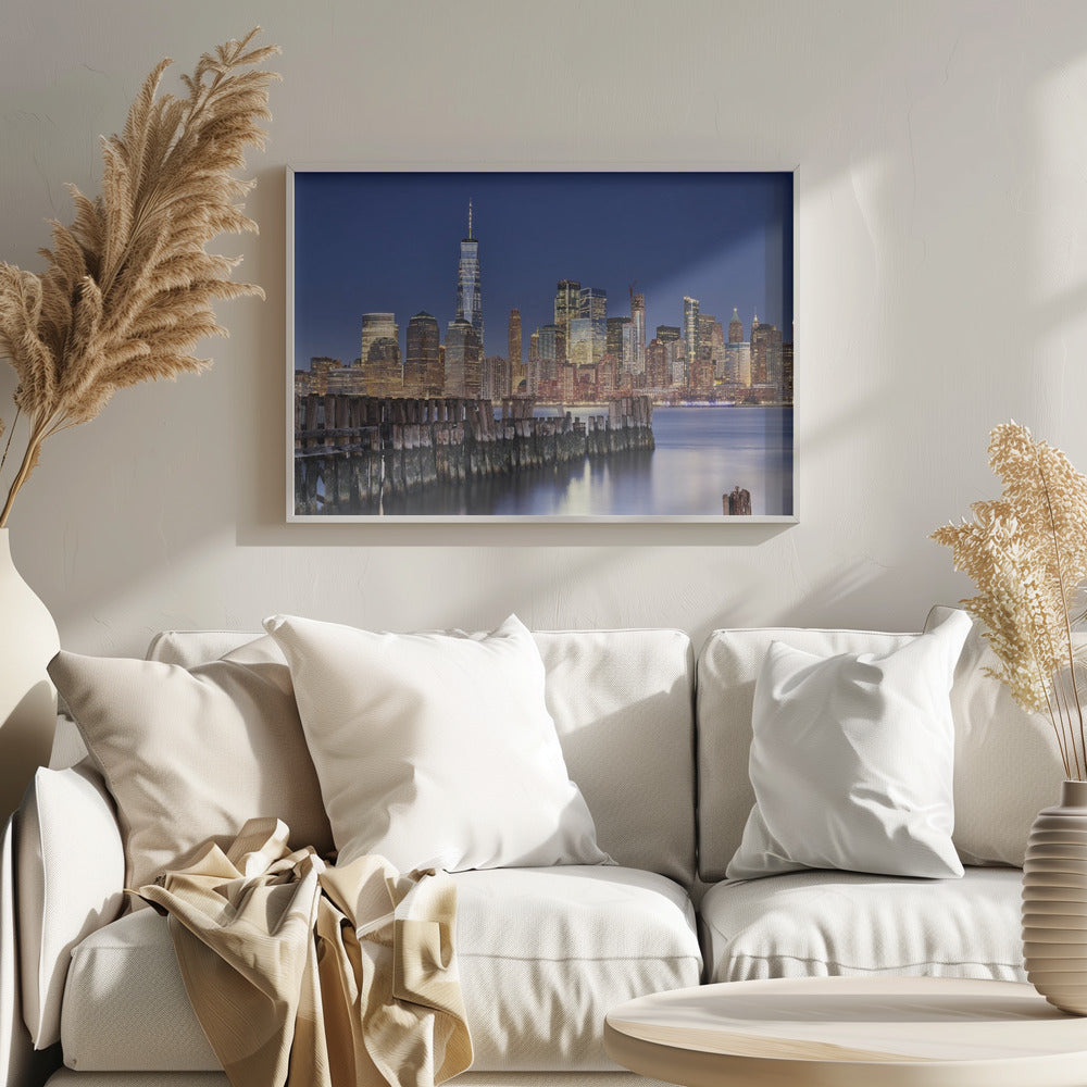 A framed photograph of the Lower Manhattan skyline at dusk. The One World Trade Center and other skyscrapers are brightly lit against a deep blue sky. In the foreground, an old wooden pier extends into the water, which reflects the city lights. Wall Art