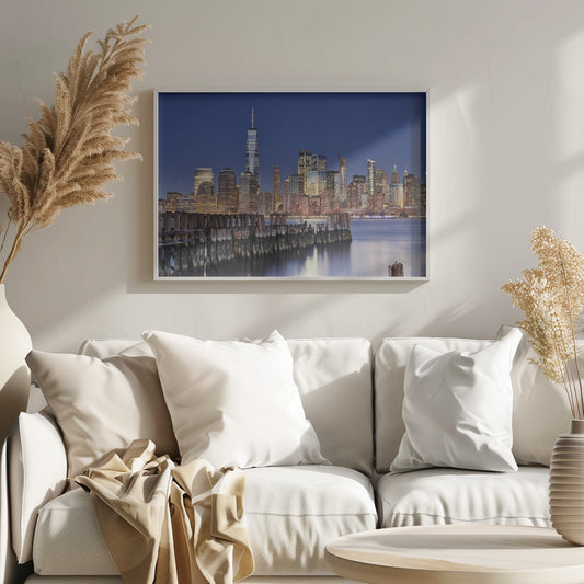 A framed photograph of the Lower Manhattan skyline at dusk. The One World Trade Center and other skyscrapers are brightly lit against a deep blue sky. In the foreground, an old wooden pier extends into the water, which reflects the city lights. Wall Art
