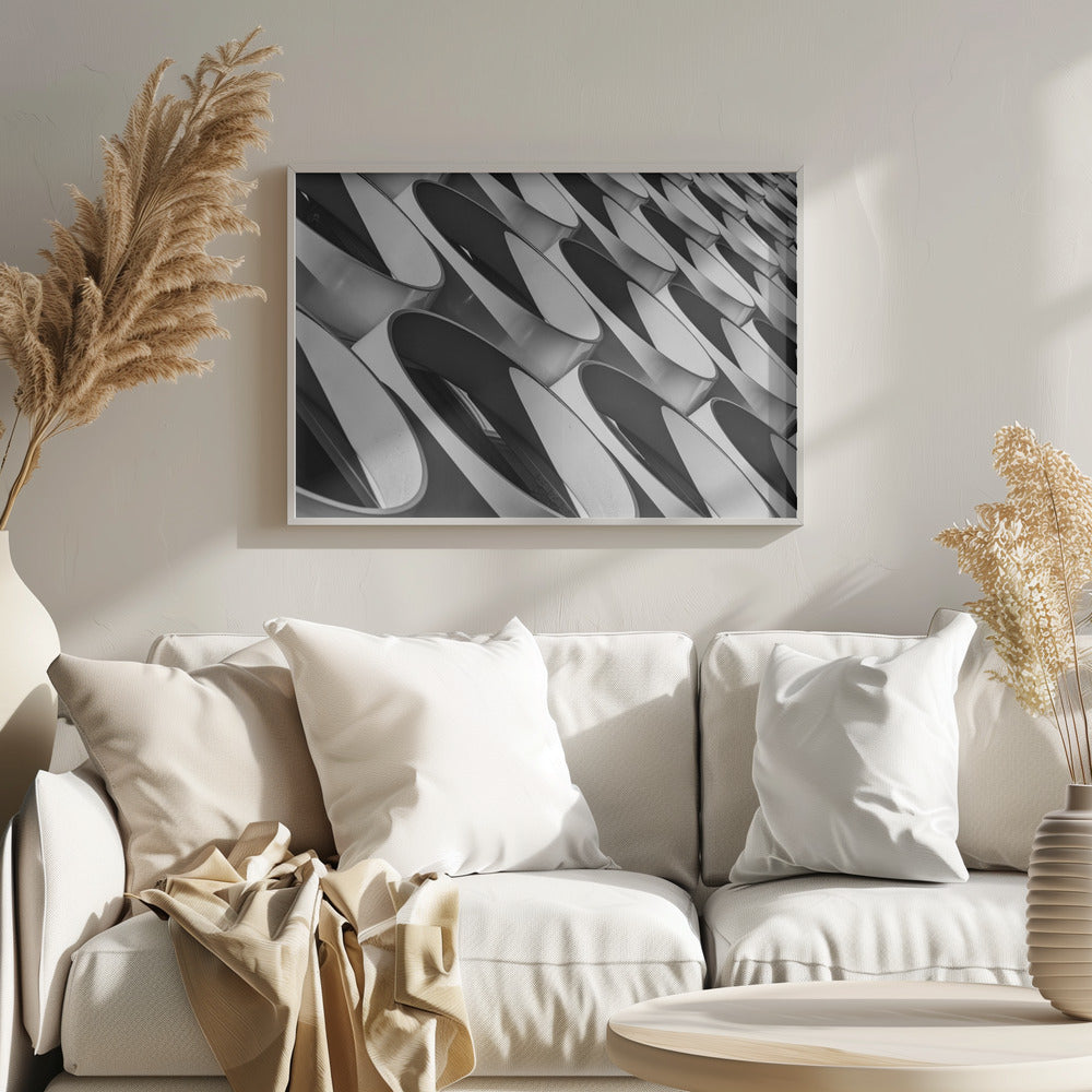 A framed, black and white photograph captures a low-angle, close-up view of a modern building's facade. The image features a repeating pattern of slanted, oval, or teardrop-shaped metallic elements, creating a dynamic interplay of light, shadow, and reflection. Print
