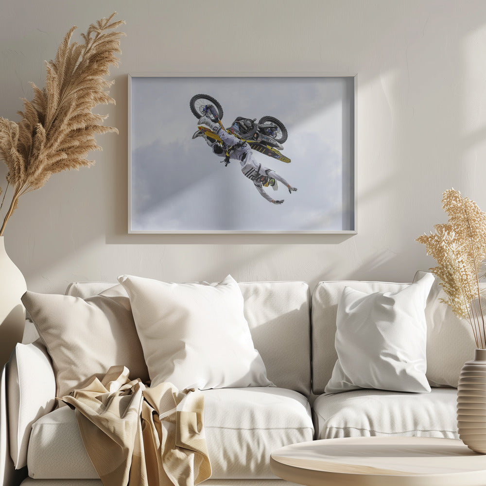 A framed action shot of a freestyle motocross rider in mid-air, performing an upside-down trick against a cloudy sky, with their body hanging below the yellow and black dirt bike. Artwork