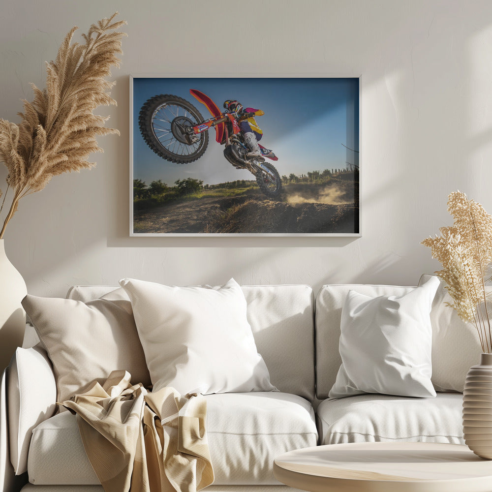 A dynamic, low-angle shot of a motocross rider in mid-air, jumping over a dirt mound. The rider is on a red, white, and blue dirt bike, wearing colorful racing gear and a helmet. The bike's front wheel is high in the air against a clear blue sky, while the back wheel is just clearing the jump, kicking up a cloud of dust. The background features a green, rural landscape. Wall Art