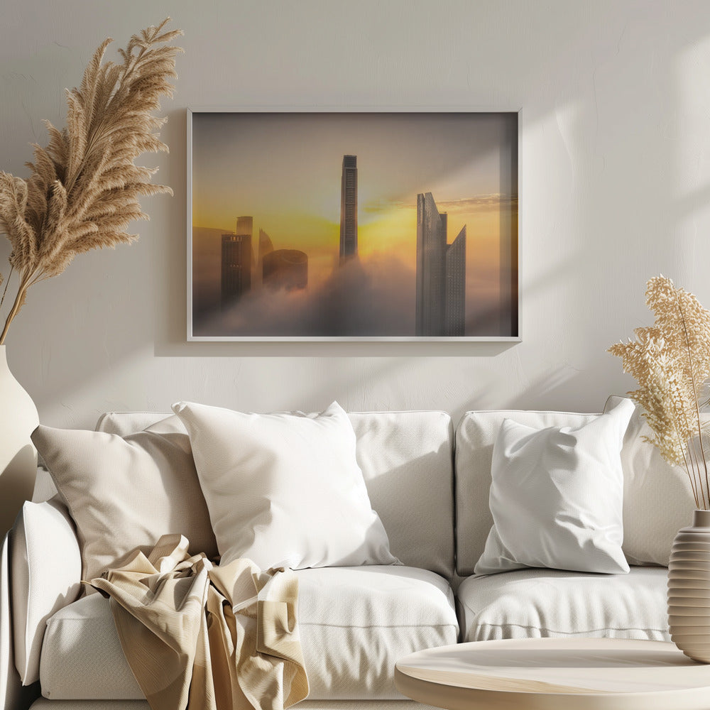 A framed photograph of a modern city skyline with skyscrapers emerging from a thick layer of clouds at sunrise, creating a dramatic and ethereal scene against a golden sky. Wall Art