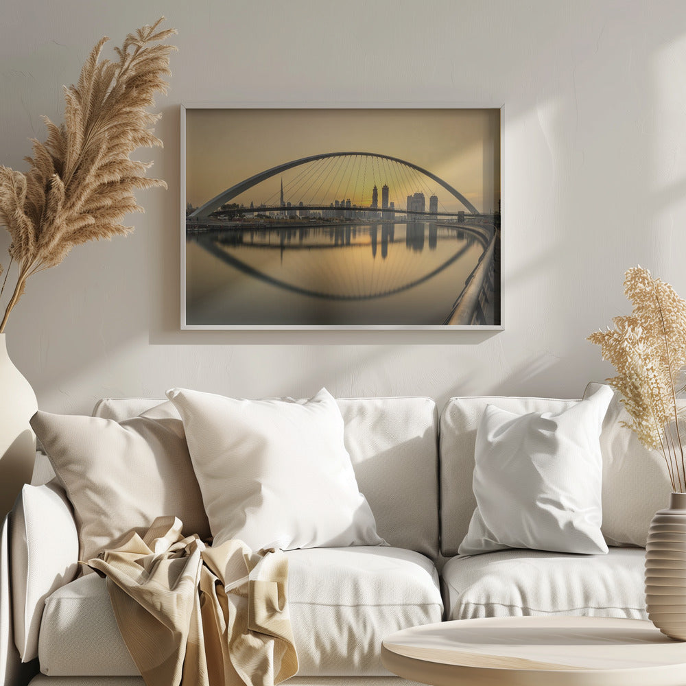 A sweeping view of the Dubai Water Canal Bridge at sunset, its white arch and cables stretching over the calm water. The city skyline, including the Burj Khalifa, is silhouetted against the golden sky, with the entire scene creating a perfect reflection in the water below. Print