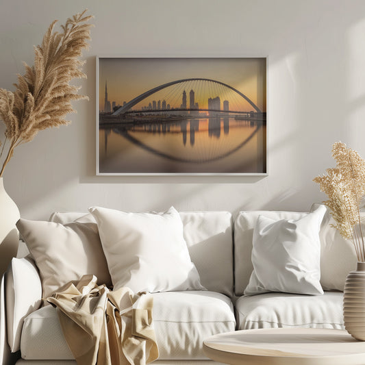 A panoramic photograph of the Dubai skyline at sunrise, viewed from across the water. A large, modern arched bridge spans the canal in the foreground, and its reflection creates a perfect oval shape in the calm water. In the background, skyscrapers, including the Burj Khalifa, are silhouetted against a glowing orange and yellow sky. The entire scene is presented within a silver picture frame. Wall Art
