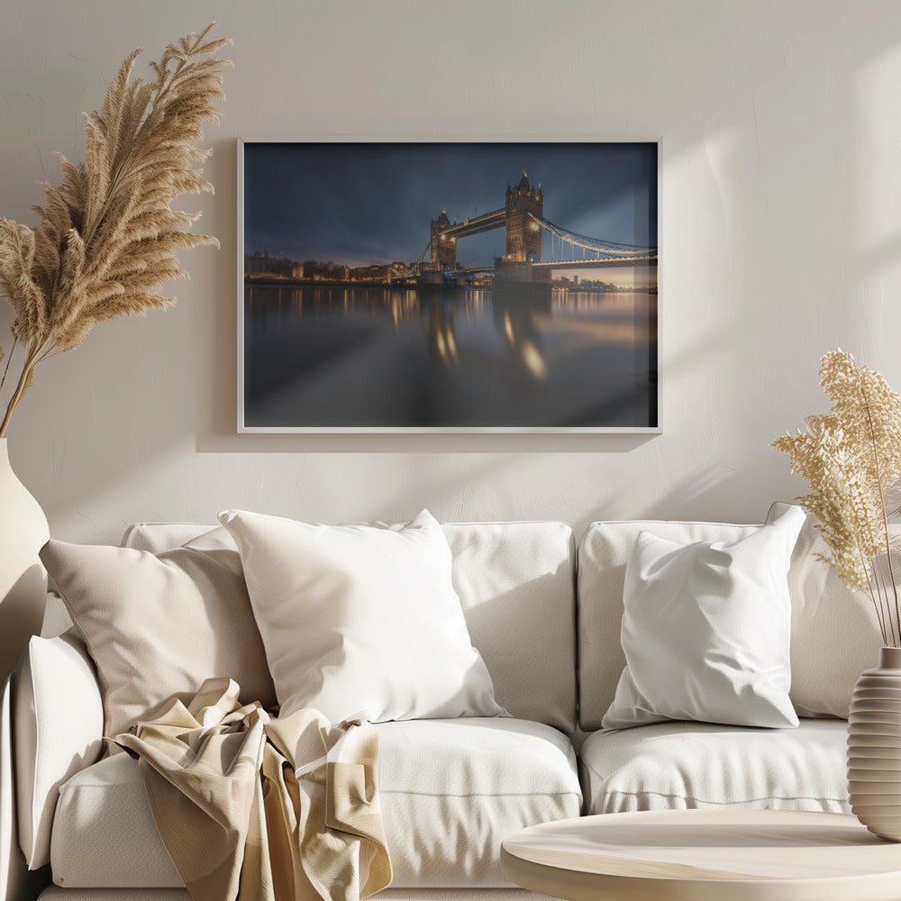 A long-exposure photograph of London's Tower Bridge at dusk, brilliantly illuminated with golden lights that cast shimmering reflections on the calm River Thames. The sky is a deep, dark blue, and the city skyline glows in the background, all enclosed within a silver frame. Print