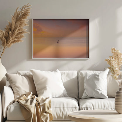 A serene photograph of a lone kayaker silhouetted against a vast expanse of calm water during a hazy, golden sunset. The sky and water are bathed in warm hues of orange, pink, and purple. A flock of birds flies in the distance, and the faint outline of a city skyline is visible on the horizon. Wall Art