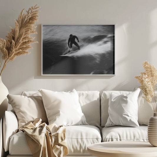 A dynamic black and white photograph of a surfer in a wetsuit carving through a wave. The image uses motion blur to emphasize the speed and energy of the water, creating a powerful and artistic effect. The artwork is presented in a simple silver frame. Print