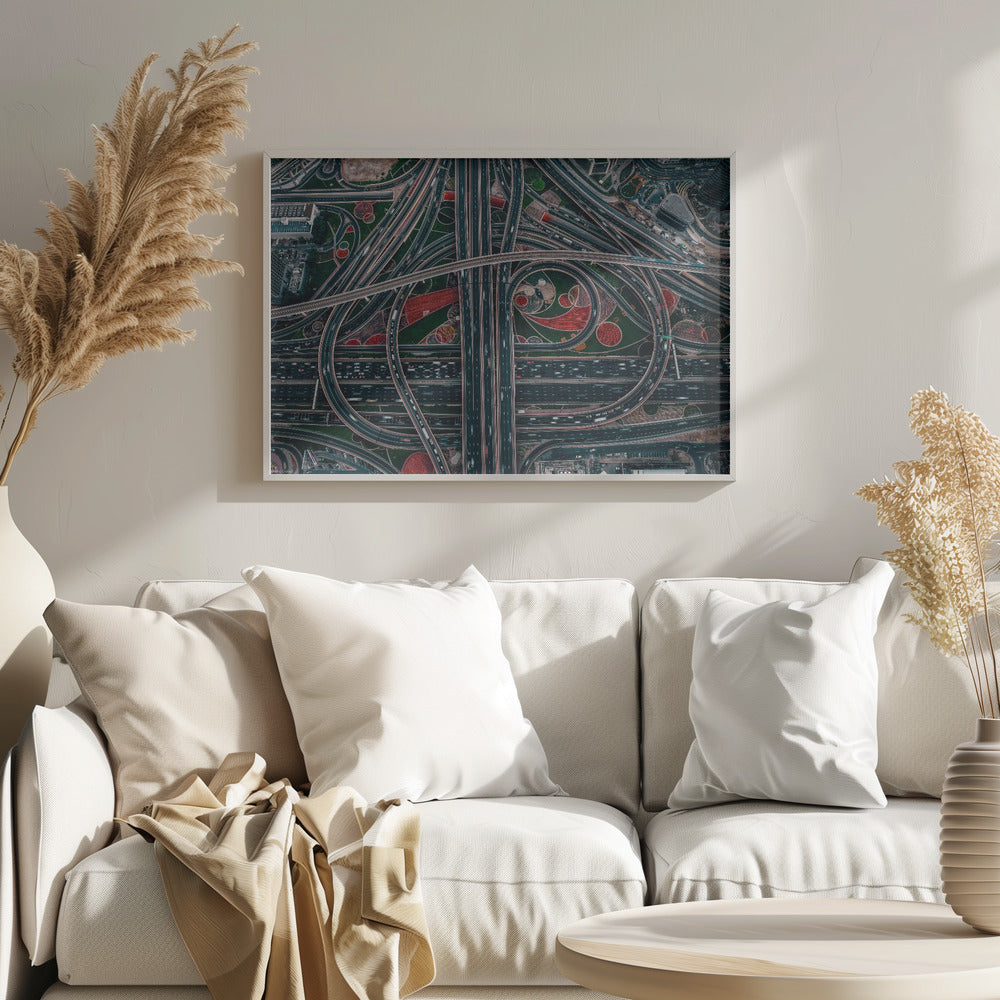 An aerial, top-down photograph of a sprawling and complex highway interchange, framed in silver. Multiple levels of roads and overpasses curve and intertwine, with blurred streaks of traffic visible. The spaces between the highways feature manicured green lawns and artistic red and patterned landscaping. Wall Art