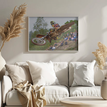 A dynamic action shot of a motocross racer in mid-air, leaning into a turn with their muddy orange dirt bike. The rider wears a colorful helmet and racing gear, with a blurred background of green trees and spectators watching from a dirt hill. The photo is enclosed in a silver frame. Decor