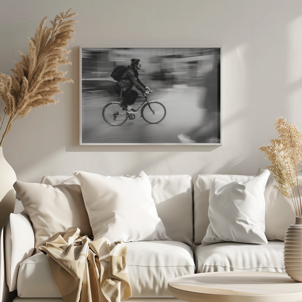 A dynamic black and white photograph capturing a person in profile as they cycle swiftly through a city. The background is a motion blur of urban scenery, highlighting the cyclist's speed. The individual wears a leather jacket, sunglasses, a backpack, and has their hair tied up. The artwork is enclosed in a silver frame. Wall Art