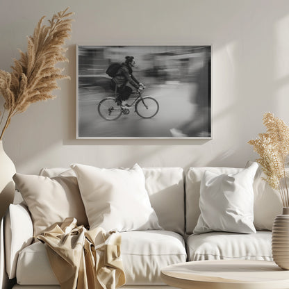 A dynamic black and white photograph capturing a person in profile as they cycle swiftly through a city. The background is a motion blur of urban scenery, highlighting the cyclist's speed. The individual wears a leather jacket, sunglasses, a backpack, and has their hair tied up. The artwork is enclosed in a silver frame. Wall Art