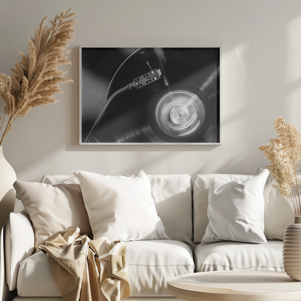 A framed, black and white, close-up photograph of a record player in action. The turntable is spinning a vinyl record, creating a motion blur on the center label. The tonearm and stylus are positioned over the grooved surface of the record. Poster