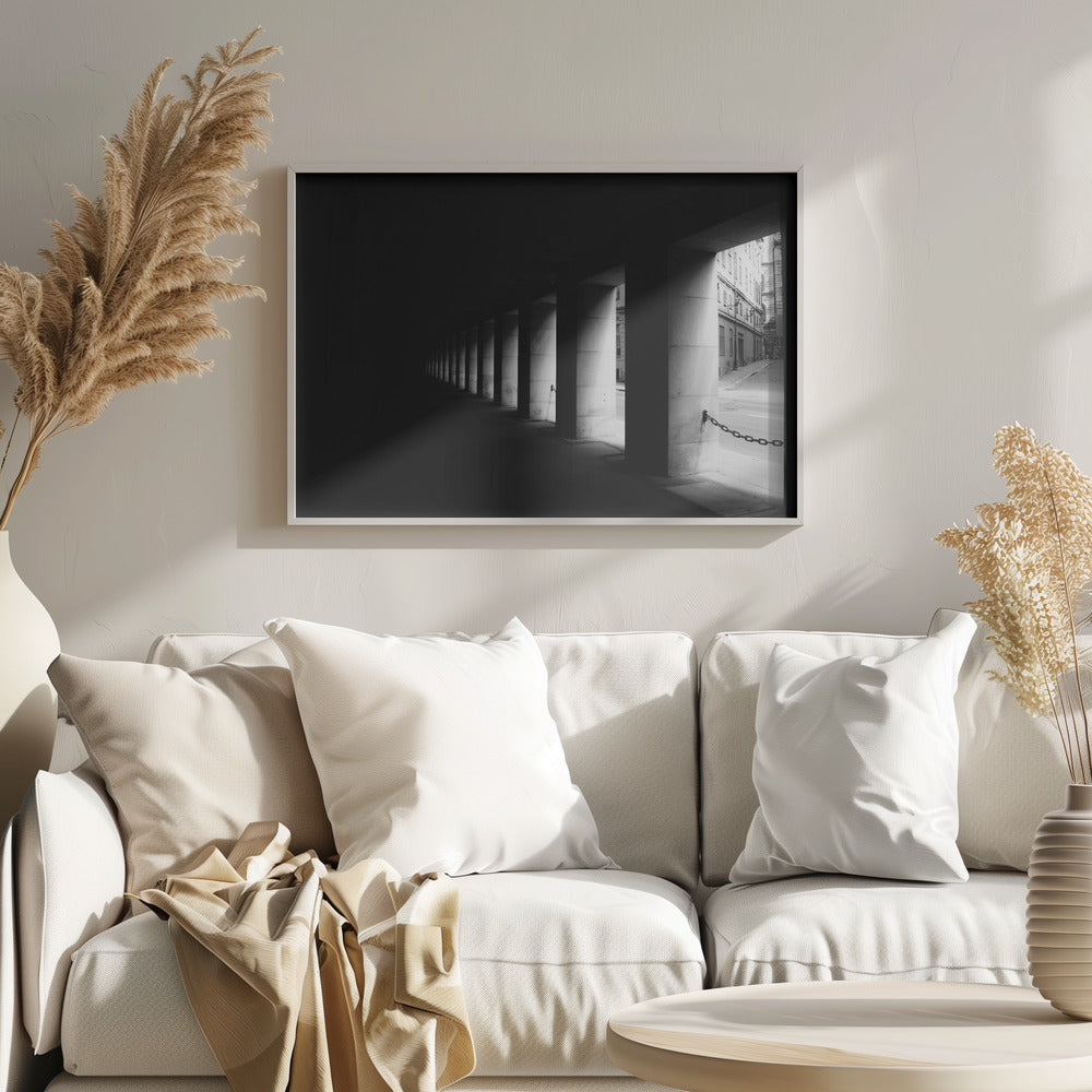A high-contrast, black and white photograph from the perspective of a dark, covered walkway lined with a row of thick pillars. Light from an adjacent street highlights the pillars, creating a dramatic play of light and shadow and a strong sense of depth. Print