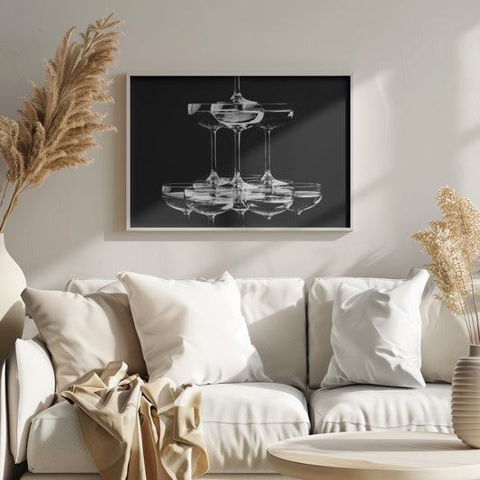 A black and white photograph of a champagne tower made of coupe glasses stacked in a pyramid shape against a solid black background, all within a silver frame. Print