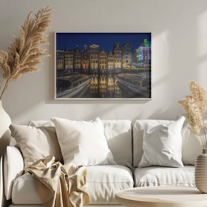 A vibrant night-time photograph of a canal in Amsterdam, flanked by two tour boats in the foreground. Traditional Dutch canal houses are brightly lit, casting warm yellow reflections on the dark water under a deep blue sky. Print