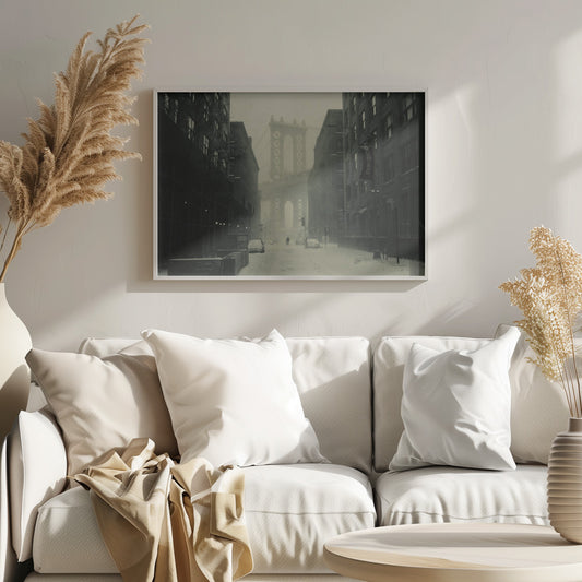 A framed, vintage-style black and white photograph capturing a snowy day in DUMBO, Brooklyn. The view looks down a snow-covered street lined with dark buildings, perfectly framing the Manhattan Bridge in the foggy distance. A few cars are parked along the street, and snow is visibly falling, creating a quiet, atmospheric winter scene. Decor