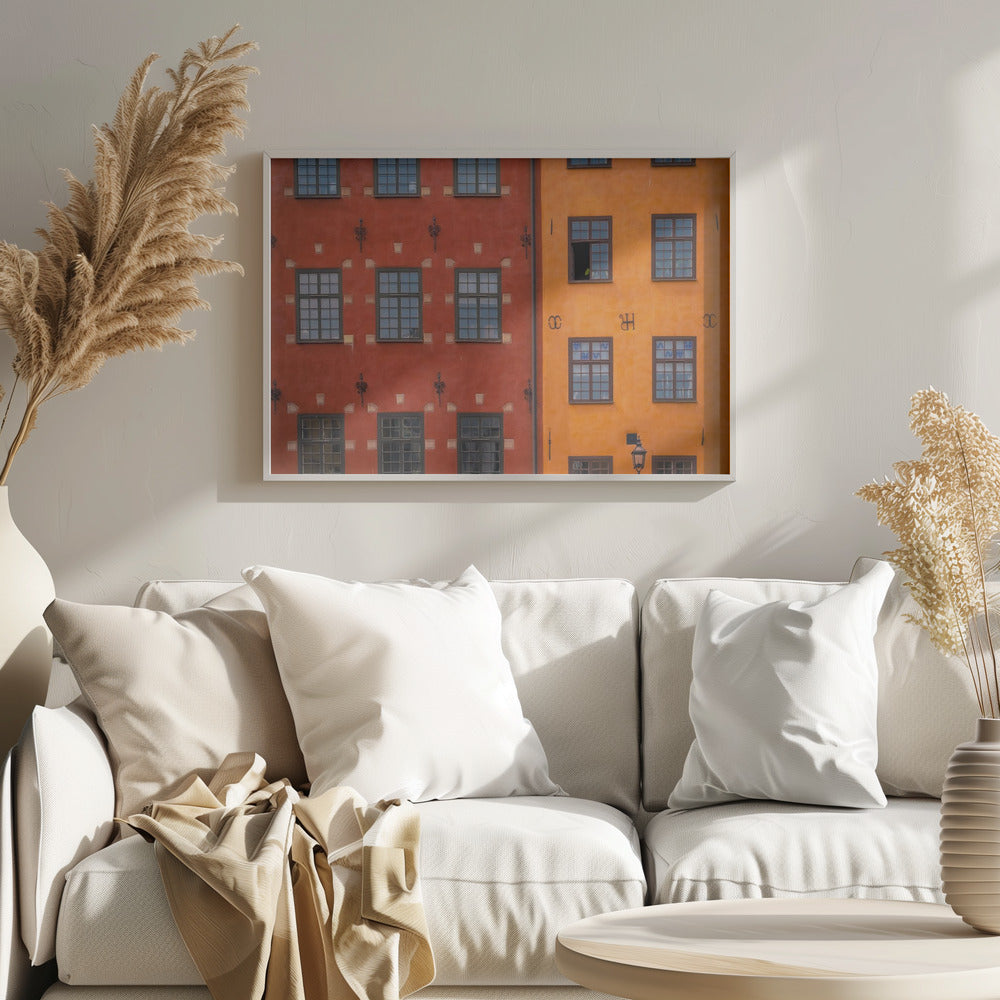 A framed close-up photograph of two adjacent, colorful building facades. The building on the left is a deep, rustic red, while the one on the right is a vibrant orange. Both feature numerous multi-paned windows with dark frames. Artwork
