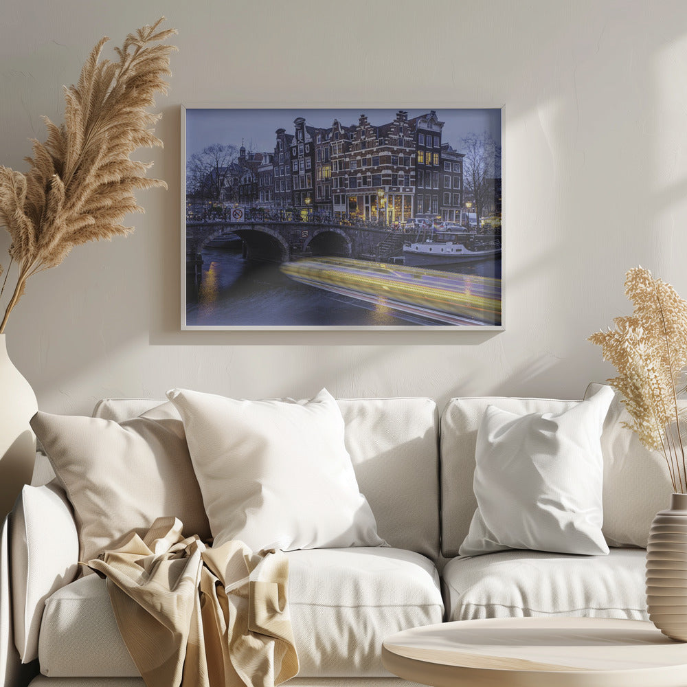 A long-exposure photograph of an Amsterdam canal at dusk, featuring a bright yellow light trail from a passing boat. In the background, illuminated historic canal houses line the water, and an arched stone bridge covered in bicycles crosses the canal. The entire image is presented in a silver frame. Print