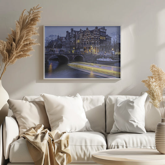 A long-exposure photograph of an Amsterdam canal at dusk, featuring a bright yellow light trail from a passing boat. In the background, illuminated historic canal houses line the water, and an arched stone bridge covered in bicycles crosses the canal. The entire image is presented in a silver frame. Print