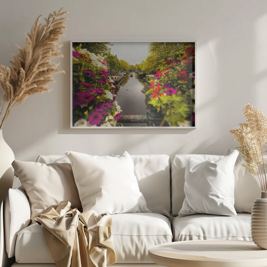 A scenic view looking down a canal in Amsterdam from a bridge, framed by vibrant, out-of-focus purple, red, and white flowers in the foreground. The canal is lined with houseboats and lush green and yellow trees, with historic buildings visible behind them. Poster