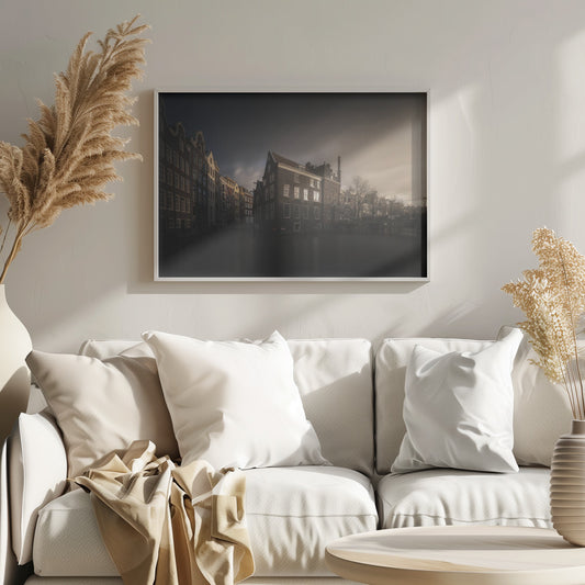 A moody, atmospheric photograph of historic canal houses in Amsterdam, viewed from the water. The scene is dark, with a dramatic, cloudy sky and a soft glow on the horizon, while some windows in the dark brick buildings are lit from within. The image is bordered by a silver frame. Wall Art