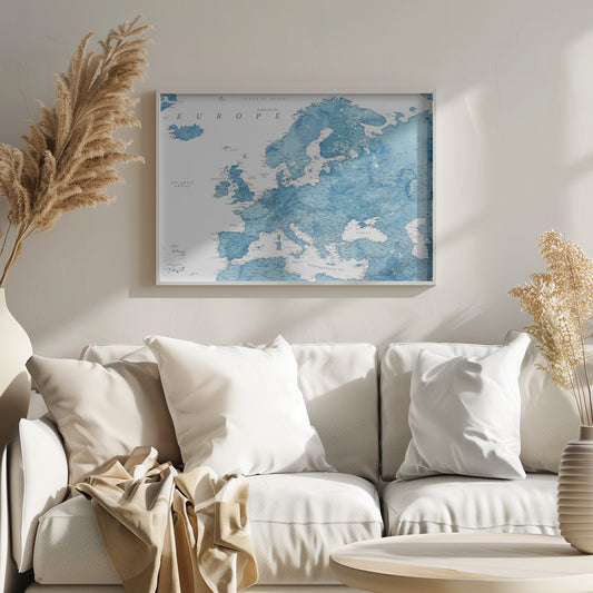 A framed map of Europe with a blue watercolor texture. The land is depicted in various shades of blue, while the oceans are white. Countries, cities, and bodies of water are labeled in black text. The map is housed in a simple, light gray frame. Poster