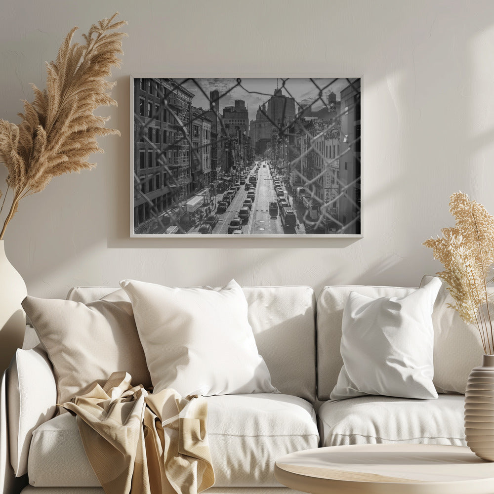 A high-angle, black and white photograph of a bustling New York City street viewed through a chain-link fence. The fence is in the foreground, framing the scene of traffic-filled streets, historic buildings with fire escapes, and skyscrapers in the distance under a cloudy sky. Wall Art