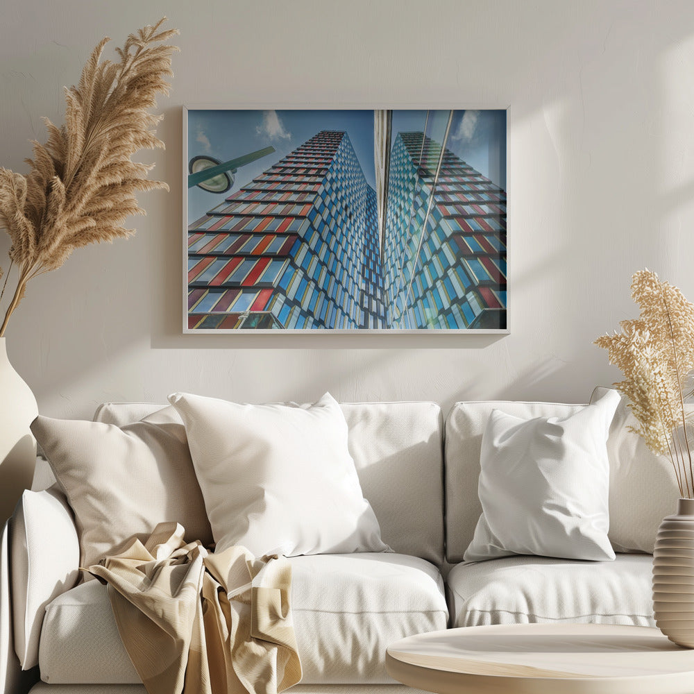 A low-angle shot of a modern skyscraper with a colorful facade of blue, red, and orange geometric panels, reflected in an adjacent glass building under a bright blue sky, all enclosed in a silver frame. Poster