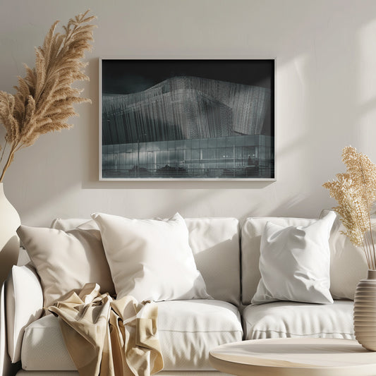 A dramatic black and white photograph of a modern building with a unique, wave-like facade made of vertical fins. The lower level is made of glass, reflecting the sky and streetlights. The entire image is presented in a silver frame. Artwork