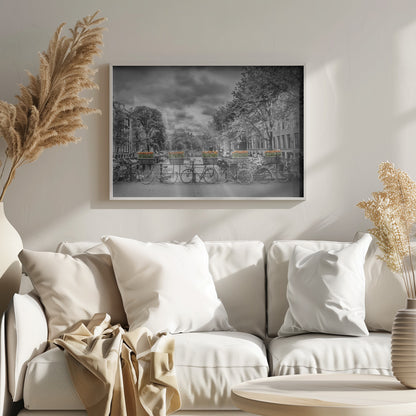 A selective color, black and white photograph of a classic Amsterdam canal scene, enclosed in a silver frame. The foreground shows a bridge lined with numerous bicycles and planters filled with vibrant orange tulips, which are the only colored elements. The background features canal houses, trees, and another bridge under a dramatic, cloudy sky with sunbeams. Print