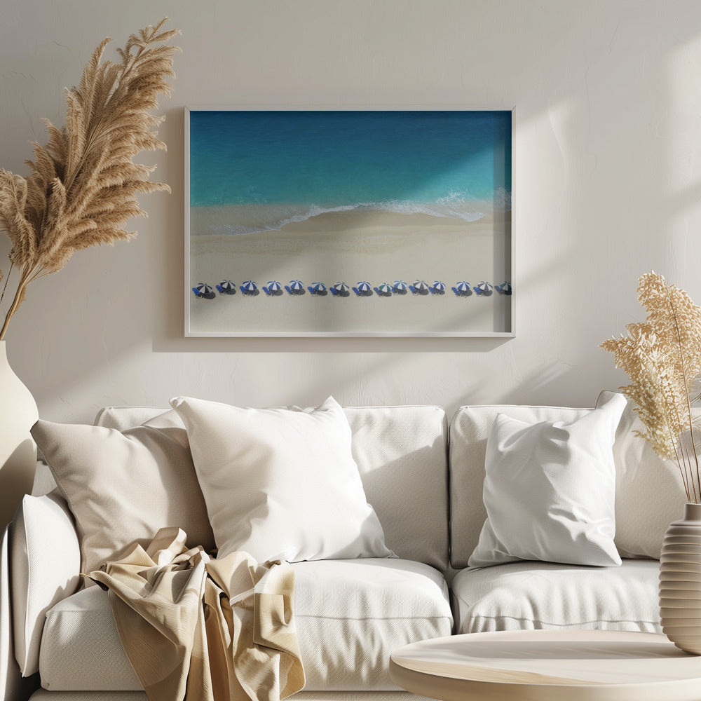 An aerial view of a pristine sandy beach with vibrant turquoise ocean waves gently lapping the shore. A long, neat row of blue and white striped beach umbrellas stands along the sand, creating a serene and orderly coastal scene, all enclosed within a silver frame. Artwork