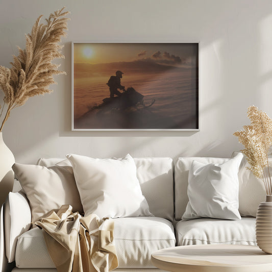 A silhouette of a person wearing a helmet and backpack stands on a snowmobile in a vast, snowy landscape during a golden sunset. The wind blows snow across the ground, creating a hazy, atmospheric effect, with mountains visible in the distance under a warm sky. Wall Art