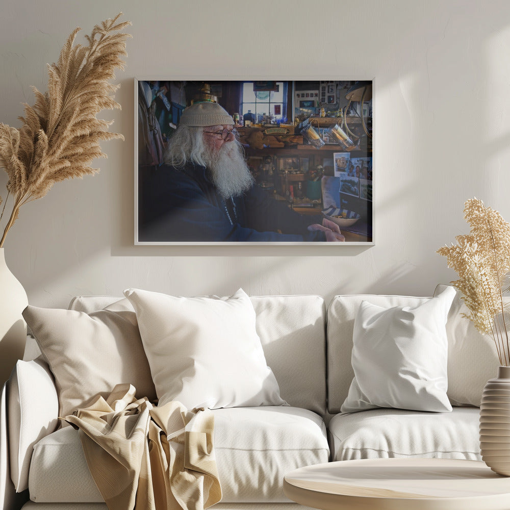 A profile portrait of an elderly man with a long, full white beard and hair, wearing glasses and a cream-colored knit beanie. He sits indoors in a warmly lit, cluttered room that looks like a cabin or a pub, with his hands clasped in front of him, gazing thoughtfully to the side. Poster