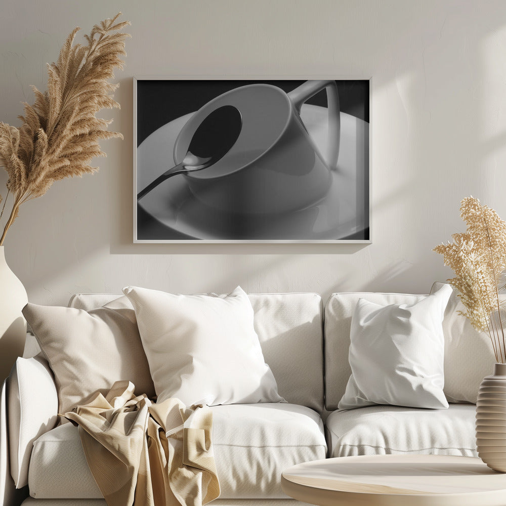 A dramatic black and white close-up photograph of a white porcelain coffee cup tilted on its saucer, with a reflective spoon resting inside. The image has high contrast against a dark background and is presented within a silver-colored frame. Decor