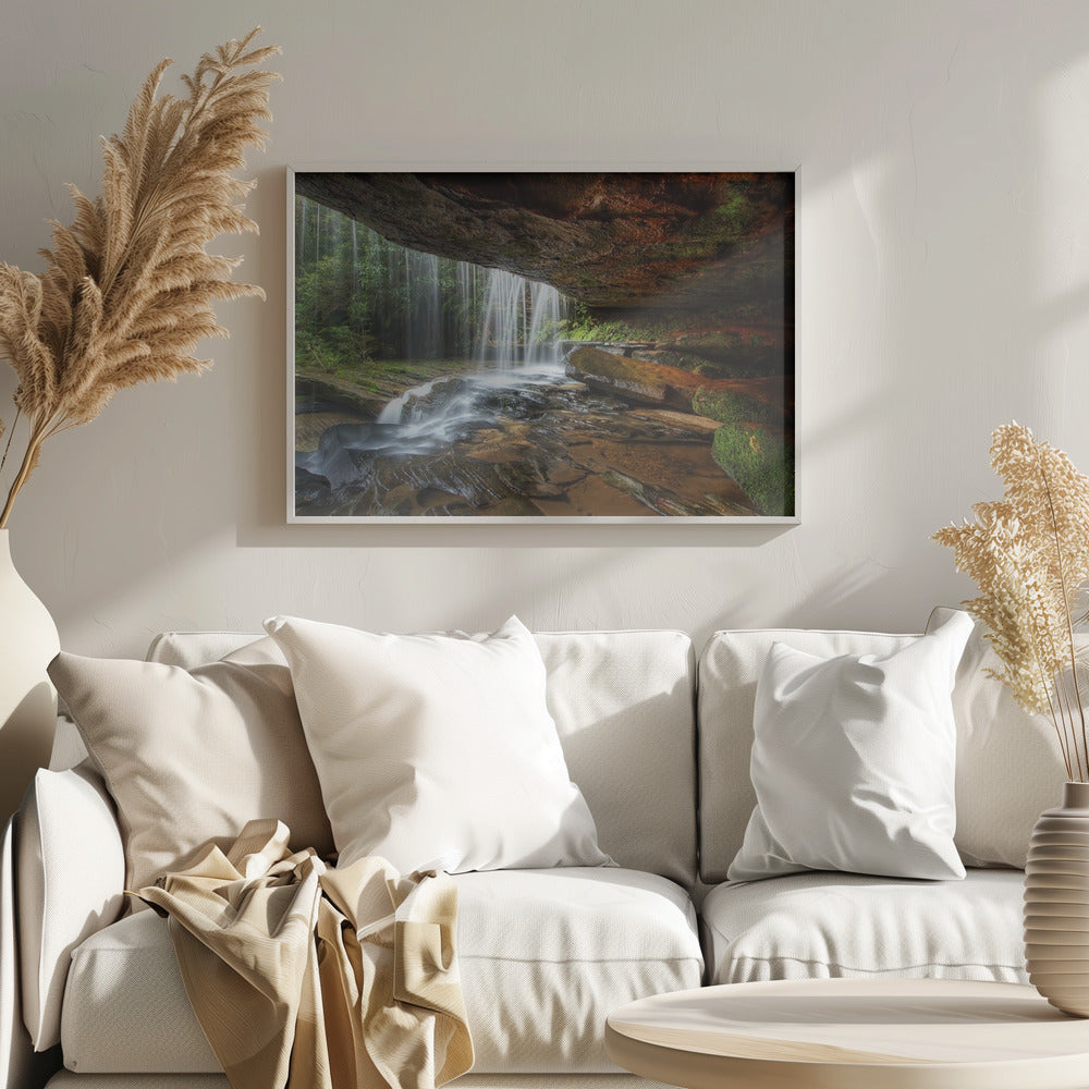 A long-exposure photograph taken from underneath a rock overhang, looking out through a cascading waterfall. The water appears silky as it flows over mossy rocks in a stream below. The scene is framed by a silver border. Print