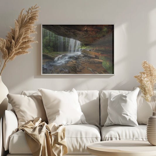 A long-exposure photograph taken from underneath a rock overhang, looking out through a cascading waterfall. The water appears silky as it flows over mossy rocks in a stream below. The scene is framed by a silver border. Print