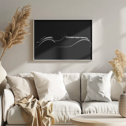 A dramatic black and white photograph showcasing the elegant silhouette of a vintage roadster. The side profile of the classic car is highlighted by subtle light, emphasizing its long hood and sweeping fenders against a solid black background, all within a brushed silver frame. Print