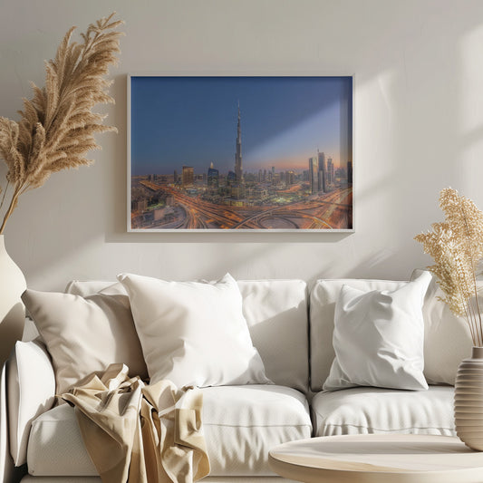 An aerial photograph of the Dubai skyline at dusk, centered on the illuminated Burj Khalifa. The sky transitions from deep blue to warm orange at the horizon, while a complex network of highways below glows with the light trails of traffic. Artwork