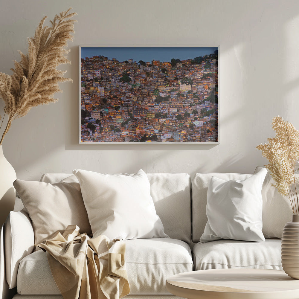 A photograph of a densely packed favela built on a hillside at dusk. The numerous small, colorful houses are illuminated from within, casting a warm orange glow that contrasts with the deep blue of the twilight sky. The image is presented within a silver picture frame. Artwork