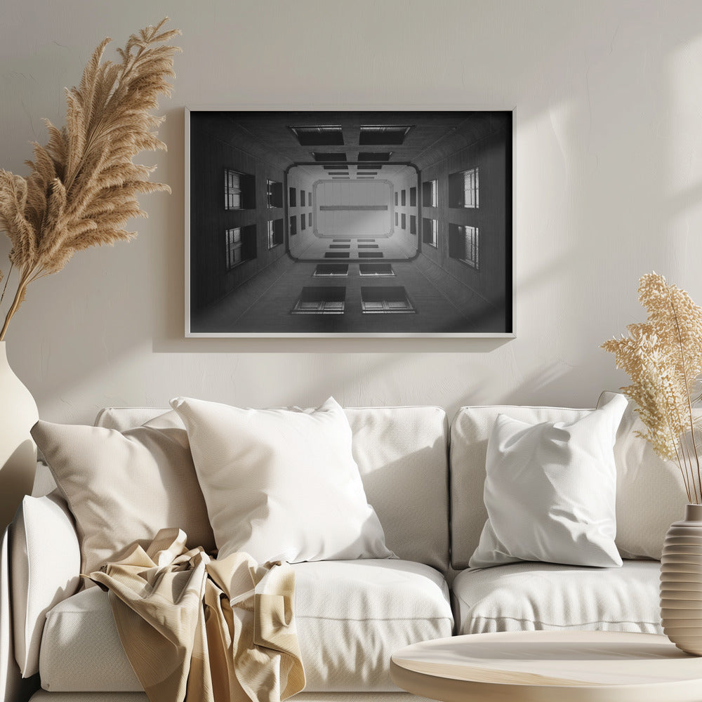 A symmetrical, black and white photograph looking directly up from the base of a building's interior courtyard. The dark walls rise up, lined with rows of windows, creating a powerful sense of perspective that draws the eye to the bright rectangle of the sky or a skylight at the top. The image is enclosed in a simple silver frame. Poster
