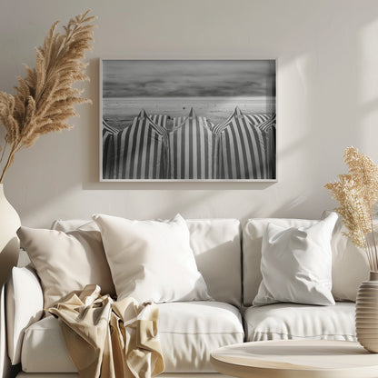 A framed, black and white photograph of striped beach tents on a cloudy day, with the ocean and distant surfers in the background. Print