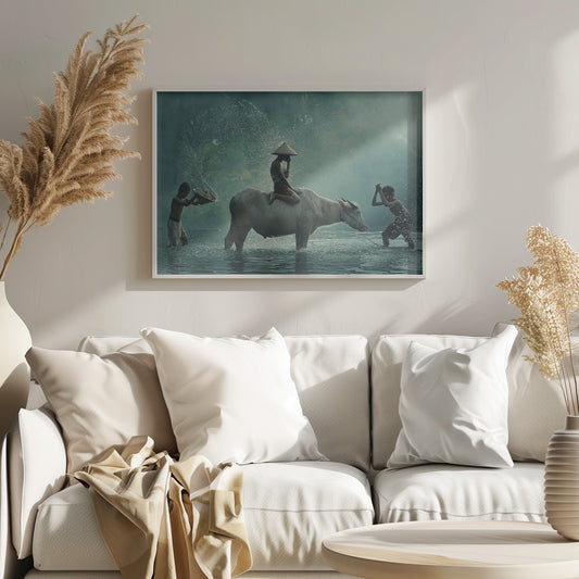 A serene photograph of three young boys playing in a river with a large water buffalo, one boy sits atop the animal while the others splash water, all set against a misty green backdrop and framed in silver. Print