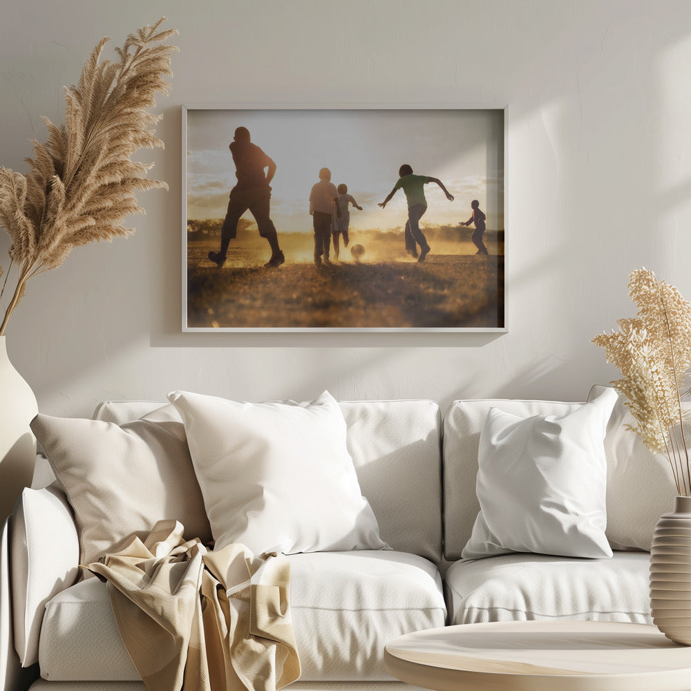A low-angle, warmly lit photograph of five children silhouetted against a bright sunset as they play soccer on a dusty field, kicking up clouds of dust in the golden light. Decor