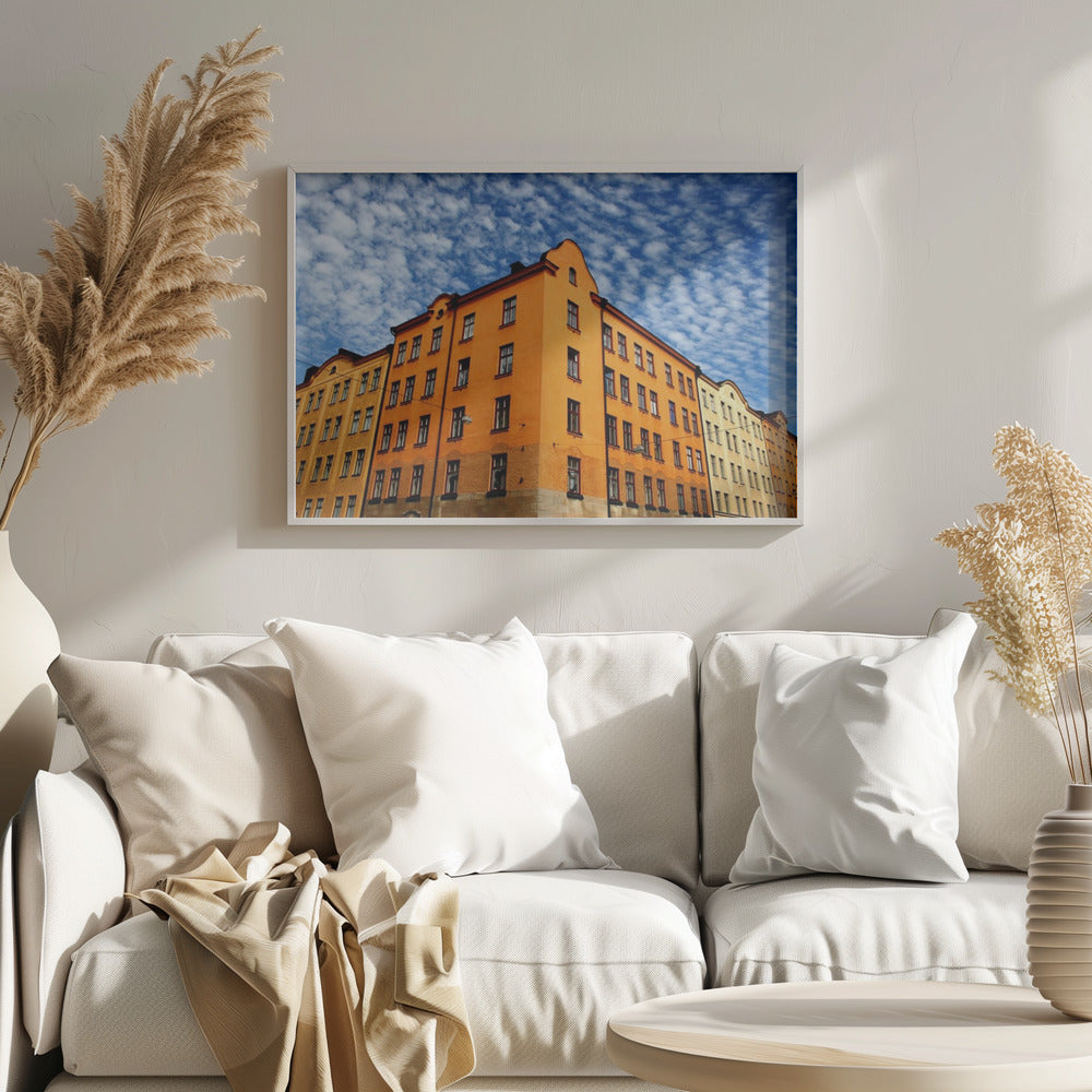 A low-angle shot of a vibrant orange apartment building against a dramatic blue sky filled with small, puffy white clouds. The building is on a corner, with another yellow-hued building visible next to it. The entire image is enclosed in a silver, brushed-metal style frame. Artwork