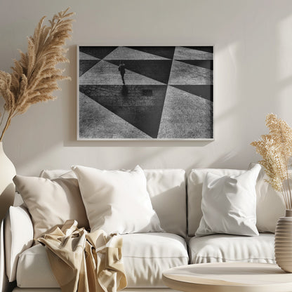 A dramatic, high-contrast black and white overhead photograph of a person walking across a paved ground with a striking geometric pattern of light and dark triangles created by strong shadows. The lone figure casts a long shadow, enhancing the minimalist and abstract composition, which is enclosed in a grey frame. Print