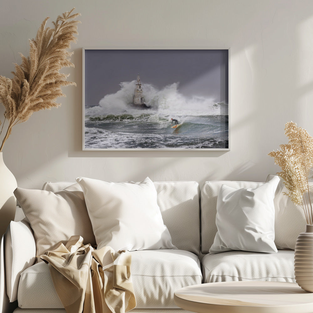 A framed photograph of a surfer in a black wetsuit riding a wave in a stormy sea. In the background, massive white waves crash against an old white lighthouse under a grey, overcast sky. Artwork