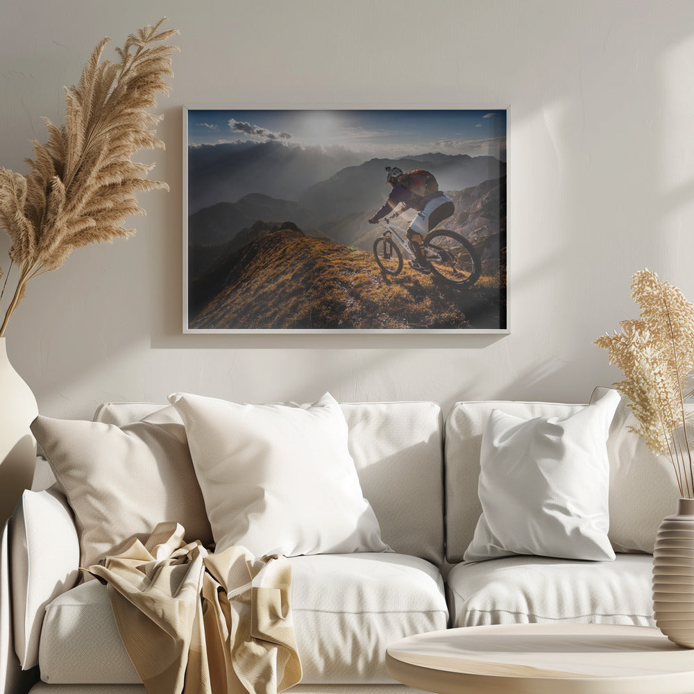 A mountain biker navigates a narrow, golden-lit ridge against a backdrop of a vast mountain range. The sun is breaking through clouds in the distance, casting dramatic rays of light across the epic landscape. Print